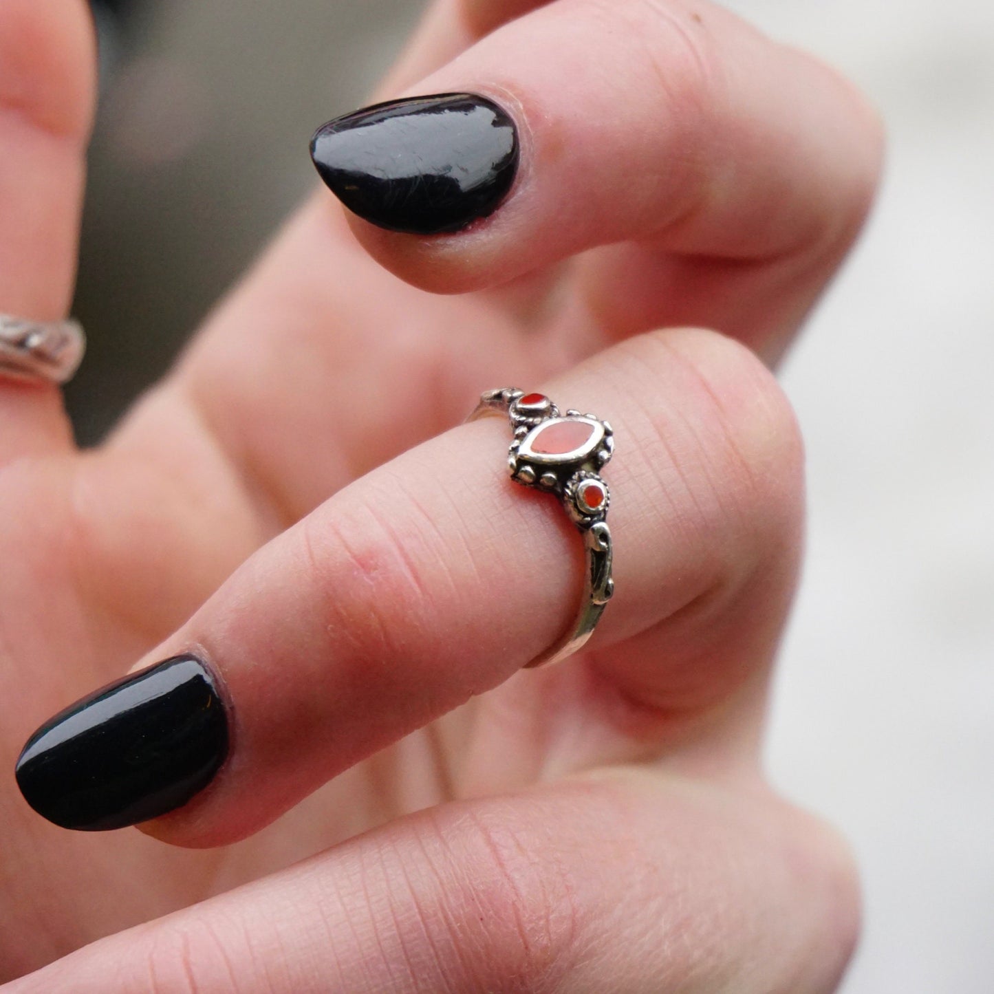 The image shows a close-up view of a hand wearing a petite vintage sterling silver ring with swirl details and small red accent stones, likely made of glass. The ring has a delicate, thin band and the red stones provide a pop of color against the dark silver metal. The person's black polished nails provide a nice contrast to showcase the intricate details of the 925 sterling silver ring.