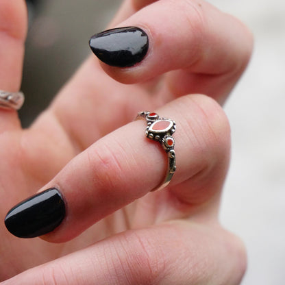 The image shows a close-up view of a hand wearing a petite vintage sterling silver ring with swirl details and small red accent stones, likely made of glass. The ring has a delicate, thin band and the red stones provide a pop of color against the dark silver metal. The person's black polished nails provide a nice contrast to showcase the intricate details of the 925 sterling silver ring.