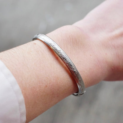 Vintage sterling silver bangle bracelet with intricate engraved design and scroll clasp, shown on wrist.