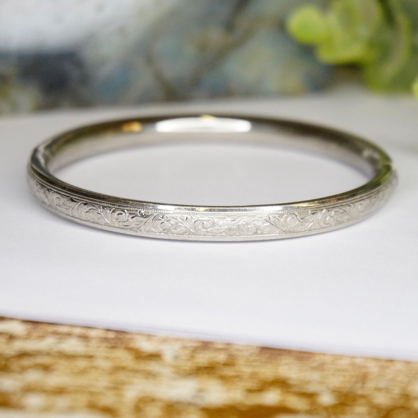 Vintage sterling silver bangle bracelet with intricate engraved floral design and scroll clasp, placed on white paper with blurred greenery in background