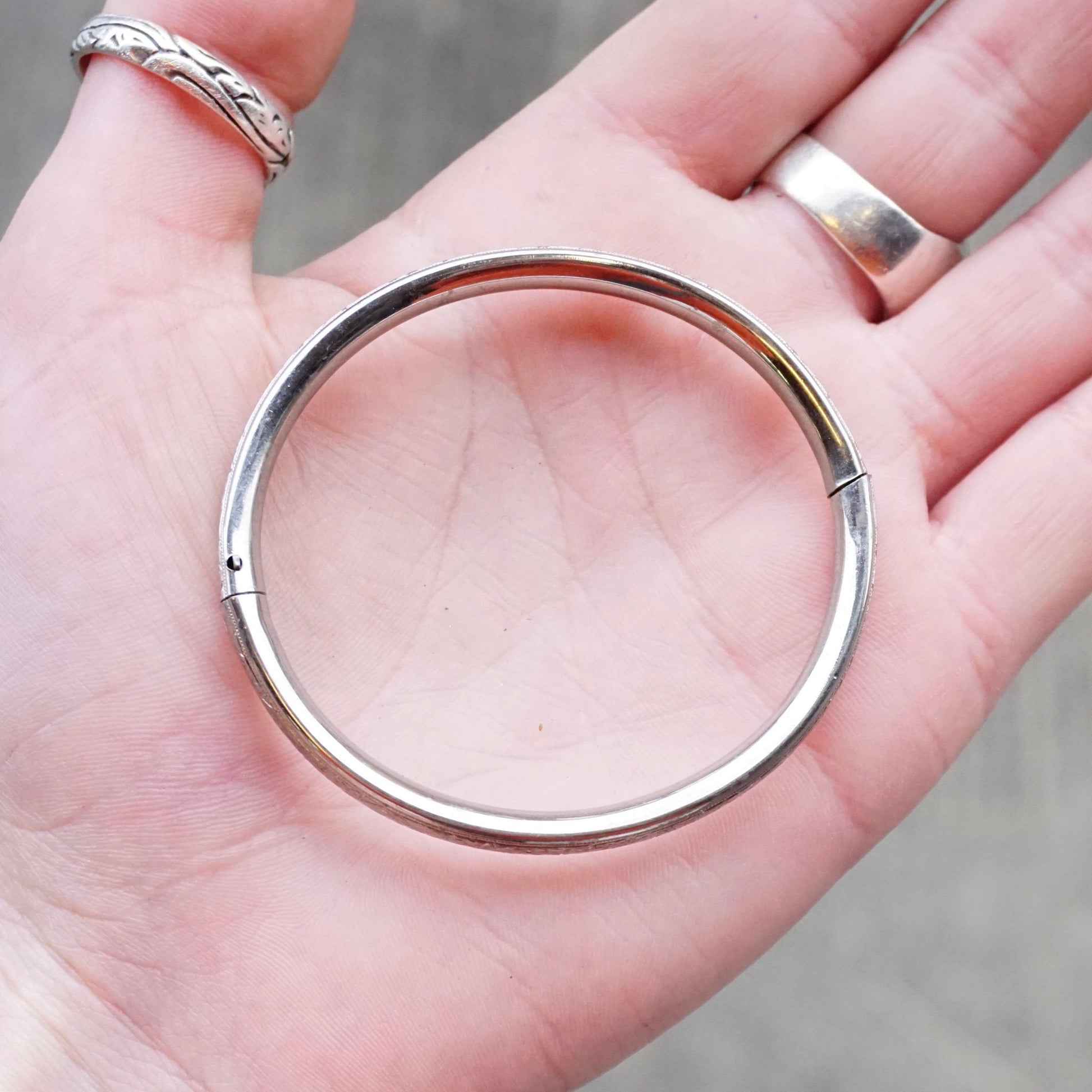 The image shows a hand holding a vintage sterling silver bangle bracelet with delicate scroll engraving details and a hinge clasp. The bangle has a bright, polished finish that gleams against the person's palm.