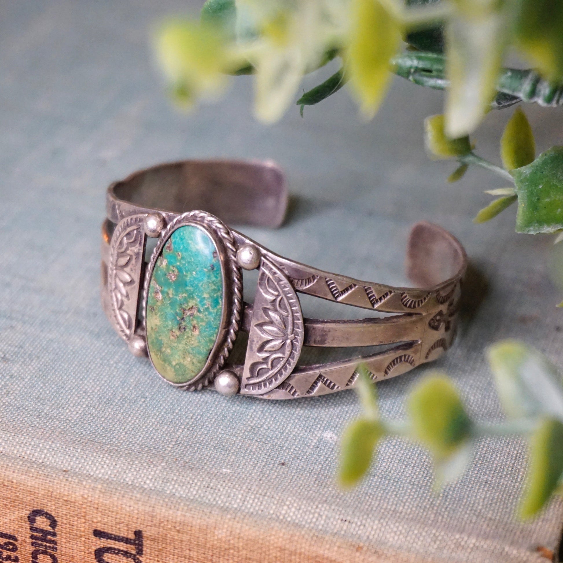 Vintage Native American coin silver cuff bracelet with natural turquoise stone, intricate handmade design, and patina, photographed on linen with sprigs of flowers.