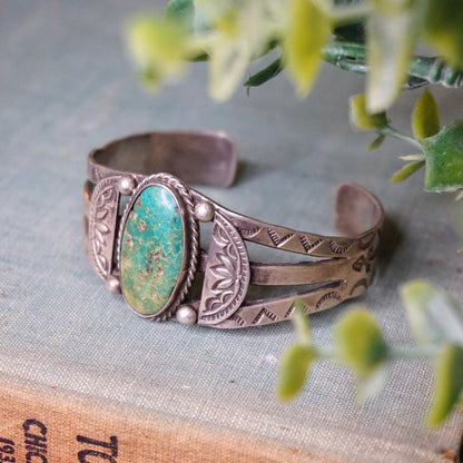 Vintage Native American coin silver cuff bracelet with natural turquoise stone, intricate handmade design, and patina, photographed on linen with sprigs of flowers.