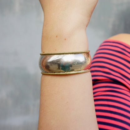 Vintage sterling silver cuff bracelet with woven gold-tone brass trim on a person's wrist, against a blurred striped shirt background.