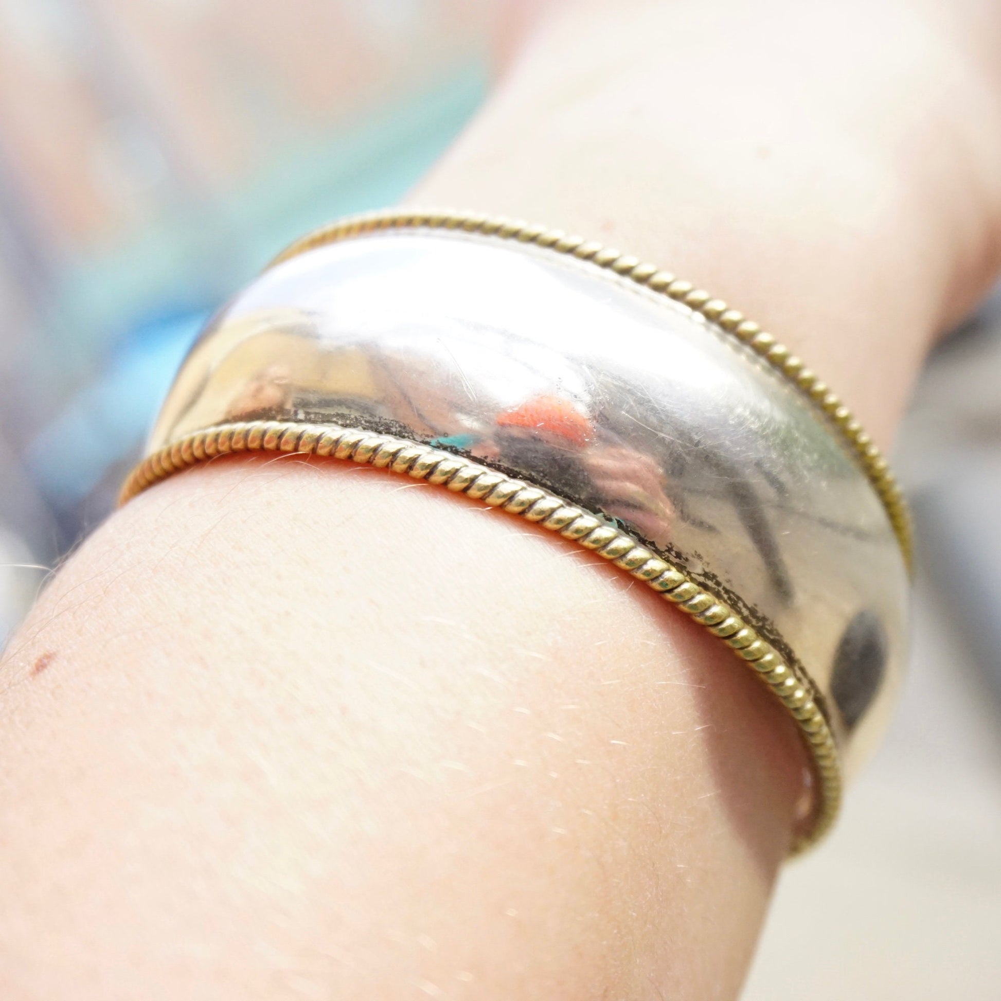 Close-up of a sterling silver cuff bracelet on a wrist, with two thin braided brass accent bands woven along the edges of the polished silver bracelet.