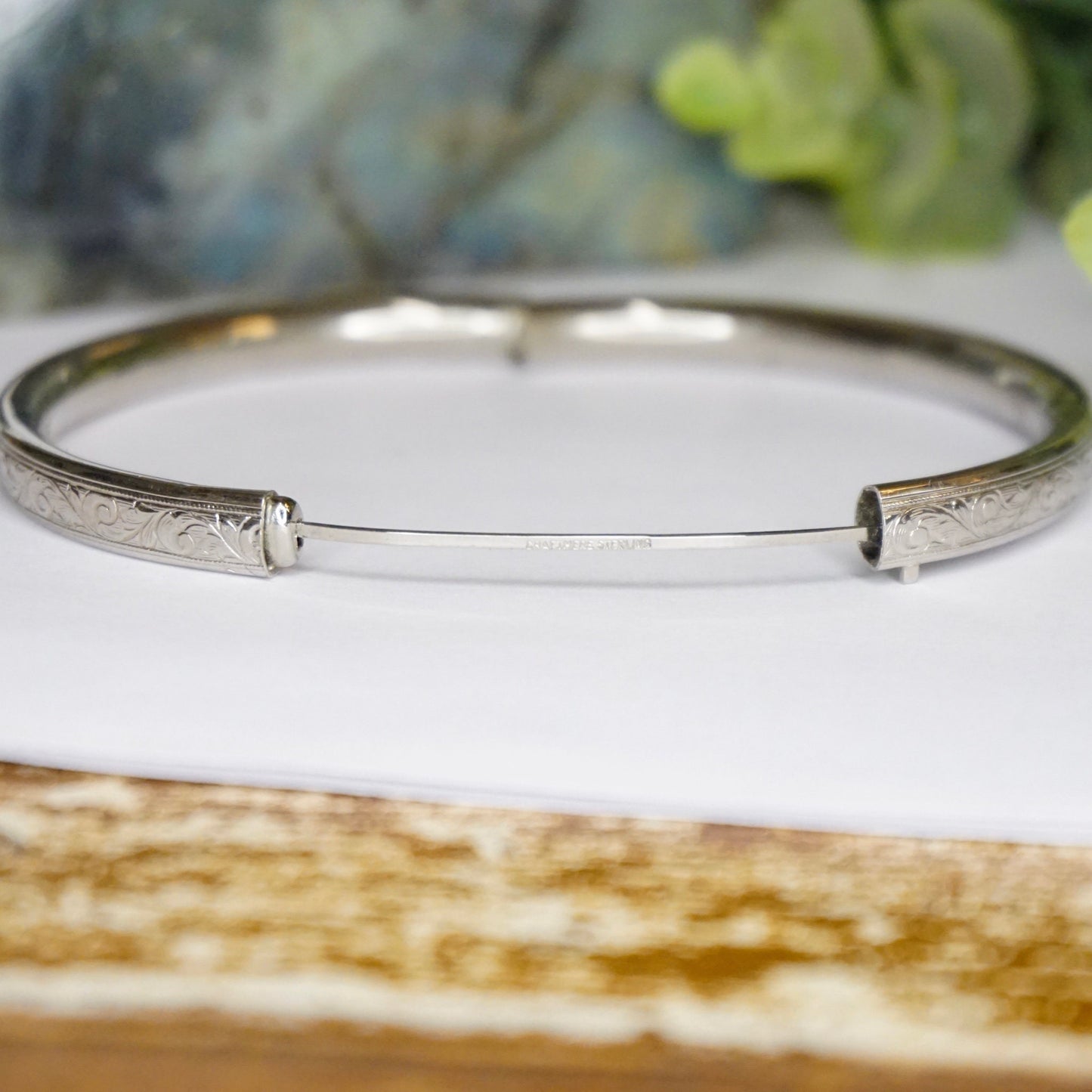 Vintage sterling silver bangle bracelet with engraved scrollwork design and secure clasp closure, displayed on weathered wood surface with succulent plant in soft focus background.