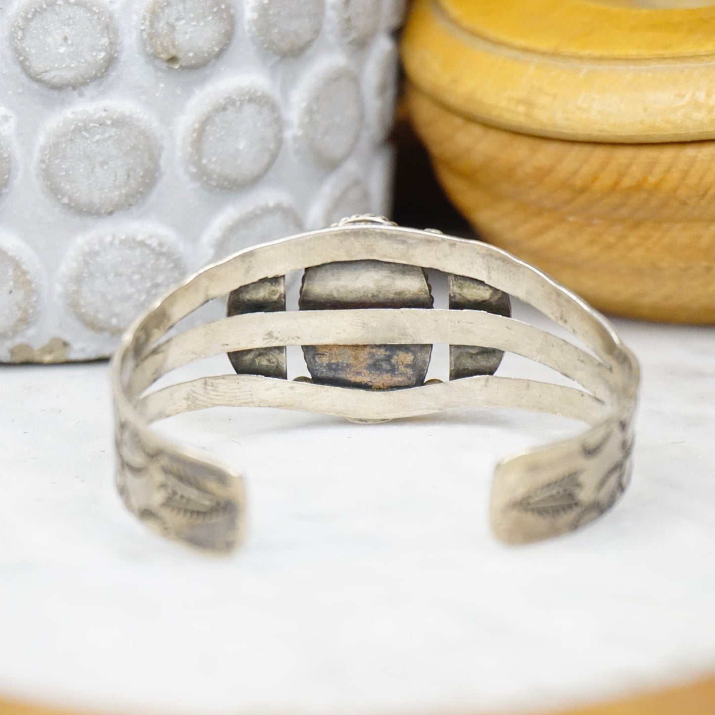 Vintage Native American coin silver cuff bracelet with intricate geometrical cut-out design and natural turquoise inlay, displayed on textured white surface with decorative ceramic jars in background, close-up view, old pawn handmade jewelry.