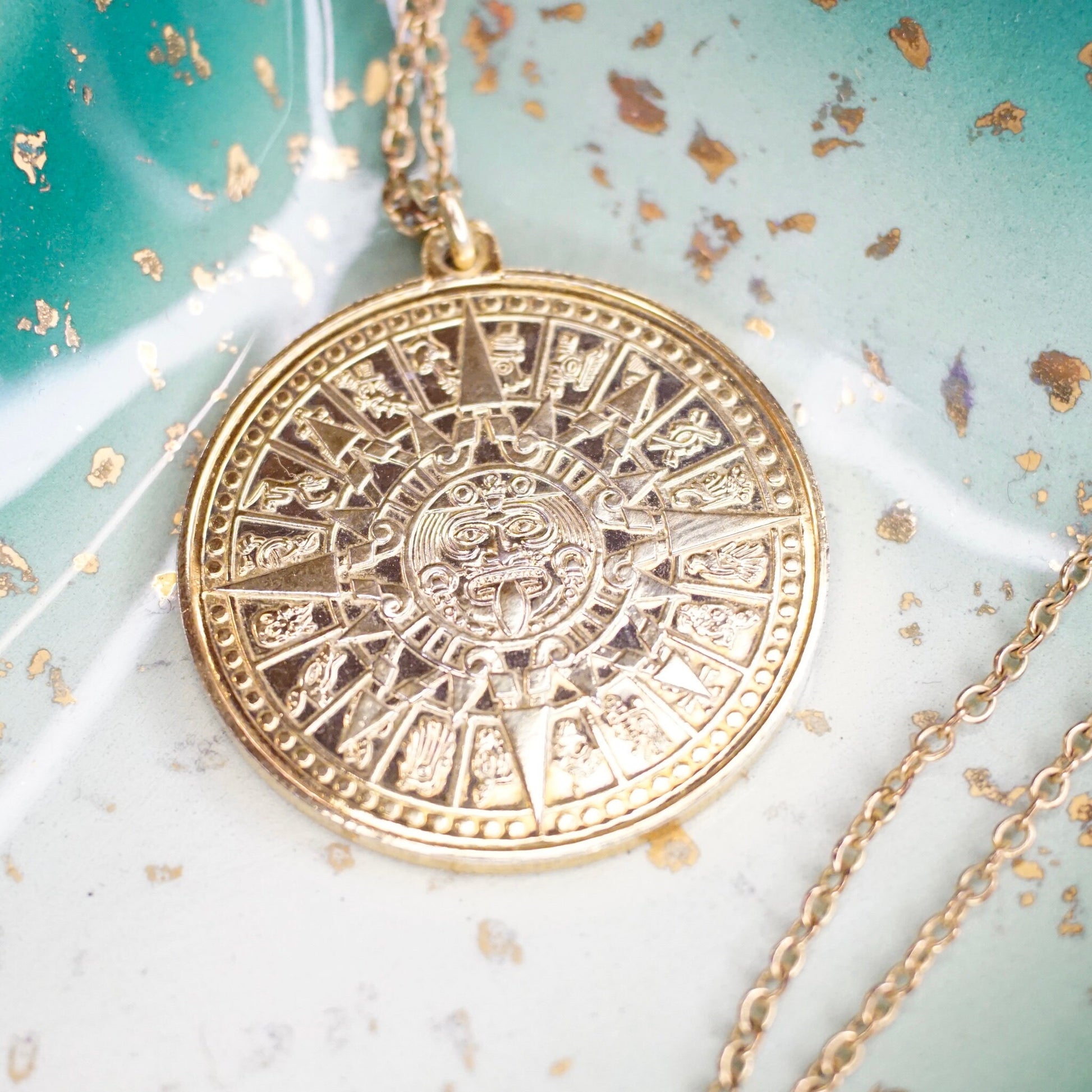 Vintage 24KT gold electroplated sterling silver Mayan sun calendar pendant necklace with double-sided Aztec sun god medallion design on a 24 inch cable chain, shown on an aged turquoise background with gold leaf flakes.