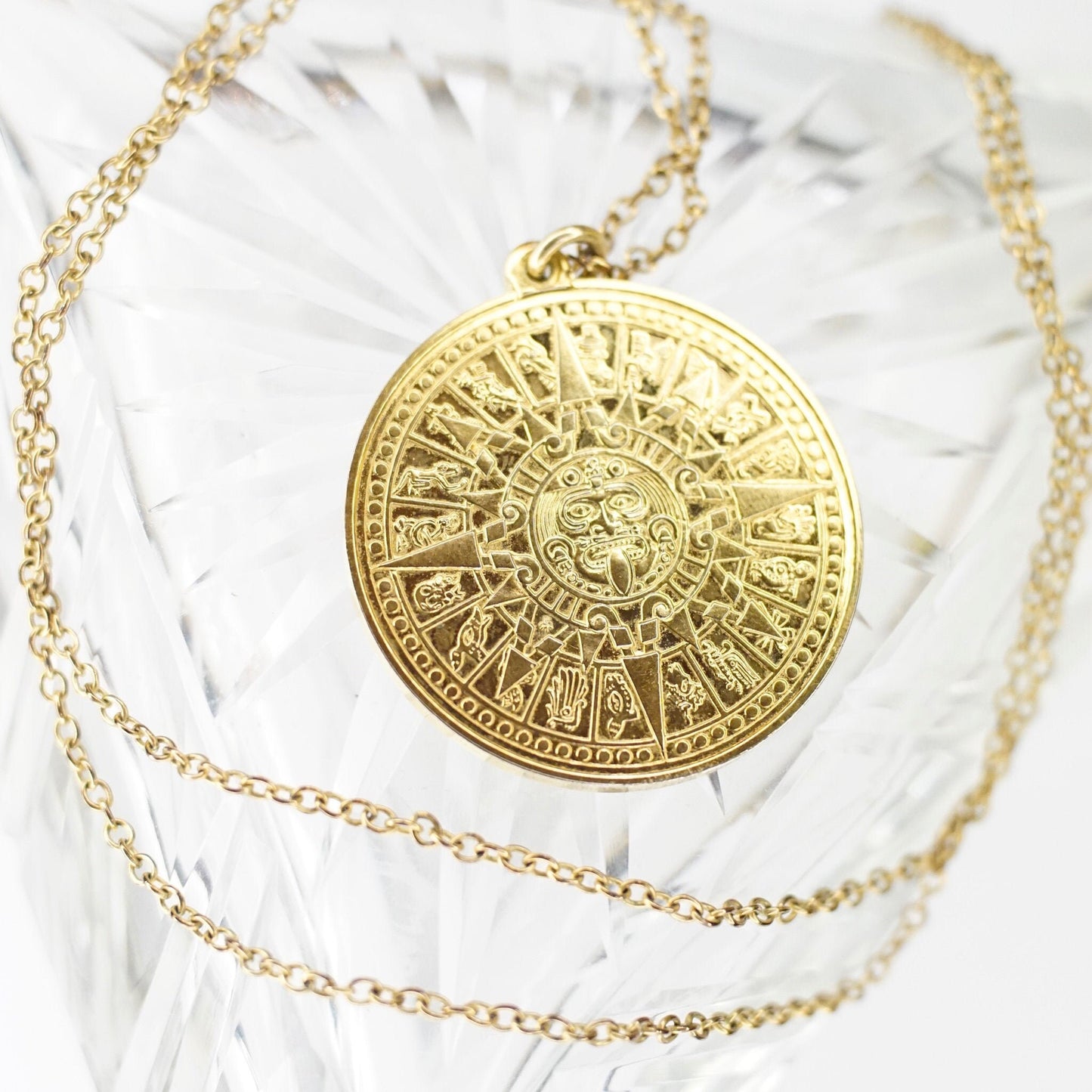 Gold Mayan calendar pendant necklace with intricate Aztec sun god design on double-sided medallion, suspended from delicate cable chain against white feather background