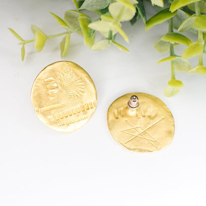 Vintage gold tone disc stud earrings with abstract hammered and stamped details, shown alongside greenery on a white background.