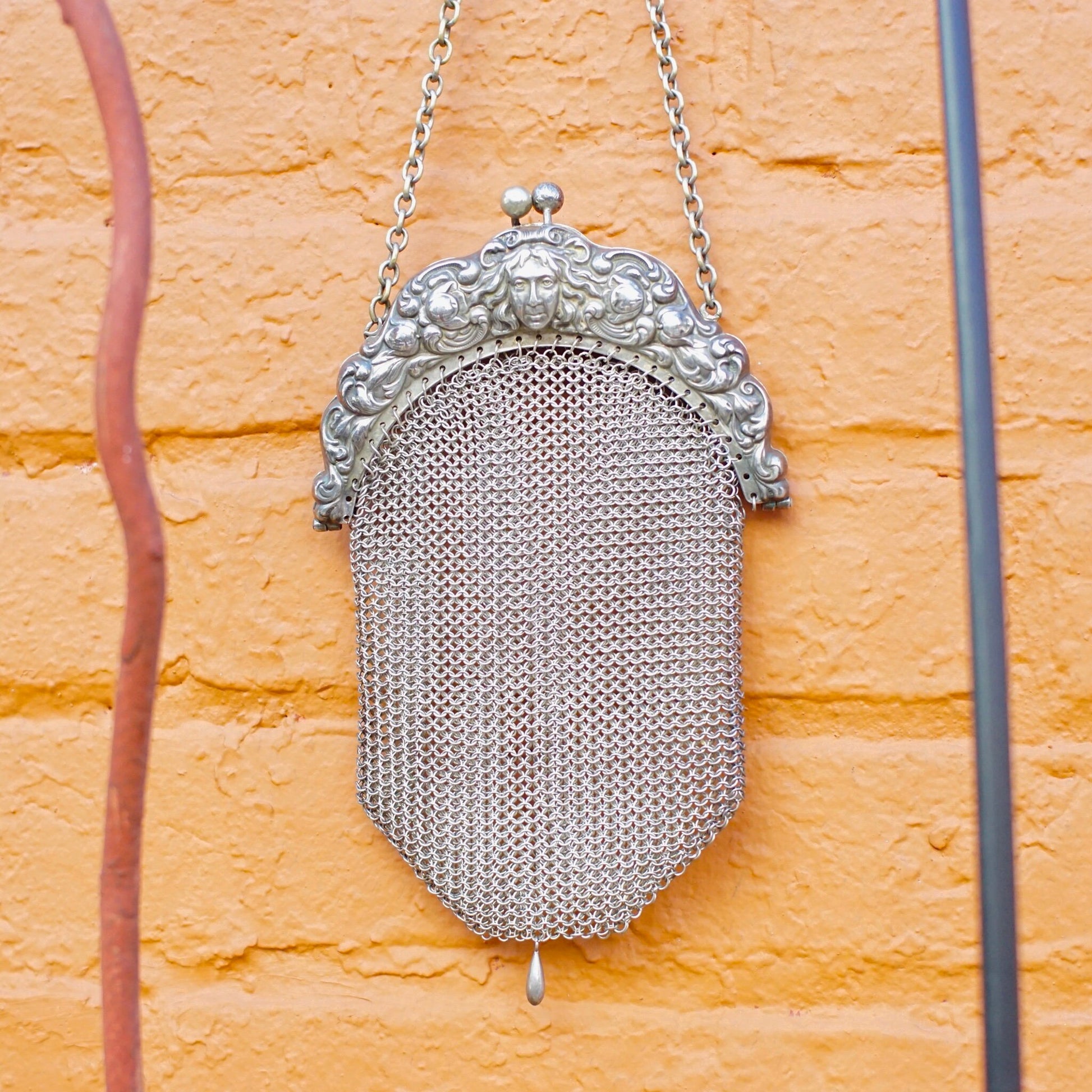 Antique Art Nouveau sterling silver chain mail purse with embossed decorative frame, hanging on an orange brick wall. The vintage mesh coin purse is in excellent condition.