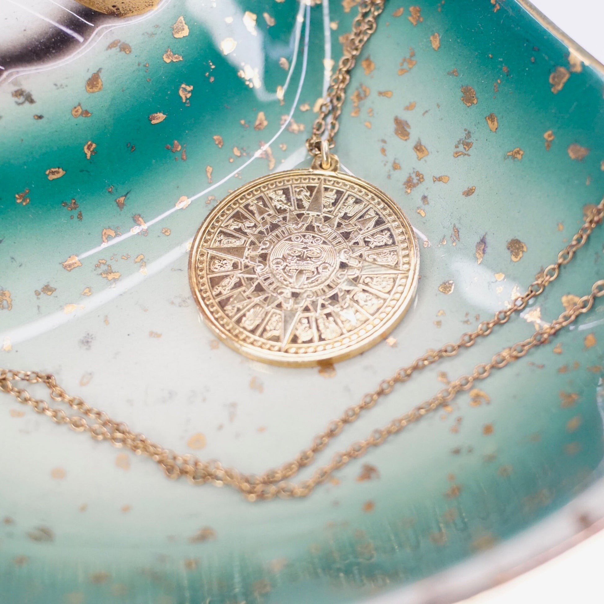 Vintage 24KT gold electroplated sterling silver Mayan sun calendar pendant necklace with ornate Aztec sun god medallion design on double-sided pendant, suspended from 24-inch cable chain, on distressed teal patina background with gold flakes.