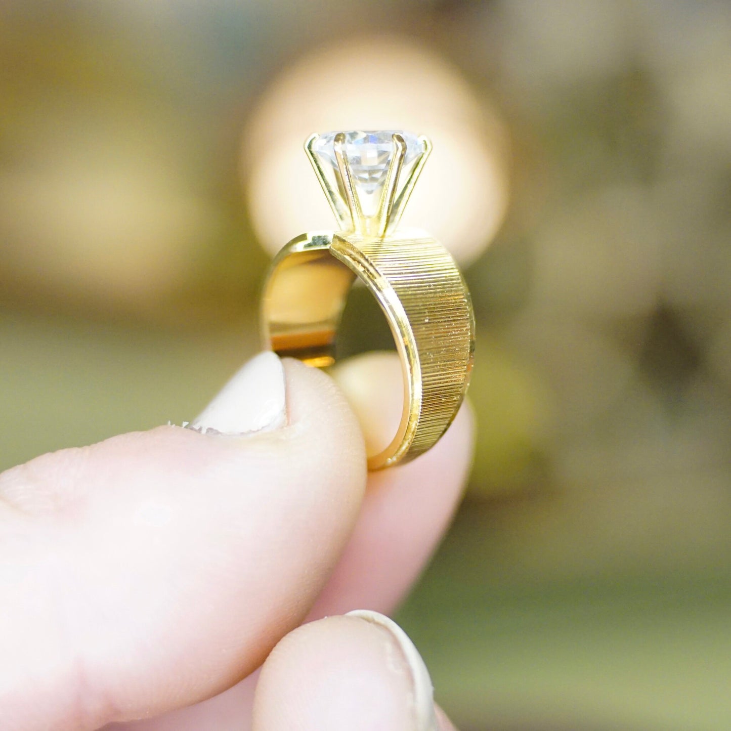 Close-up of a hand wearing a vintage gold-tone ring with a round-cut cubic zirconia solitaire set in a 6-prong setting, against a blurred natural background.