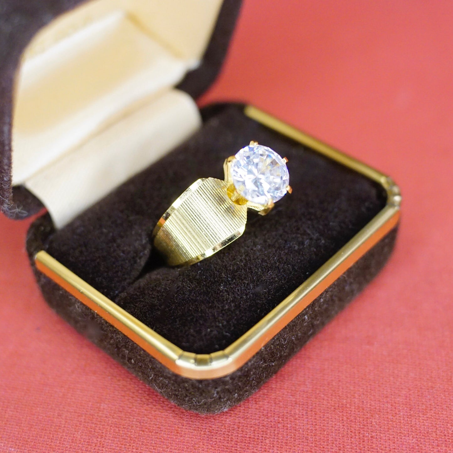 Vintage gold tone solitaire ring with round cut cubic zirconia stone in 6-prong setting, presented in jewelry box against red background.