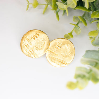 Vintage gold tone disc stud earrings with hammered and stamped abstract details, artisan gold post earrings, shown on white background with green foliage accents.