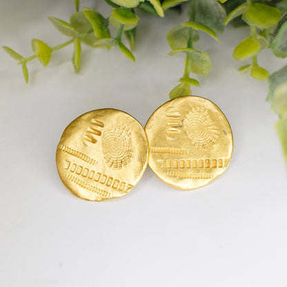 Vintage gold tone round disc stud earrings with hammered and stamped geometric details on green foliage background