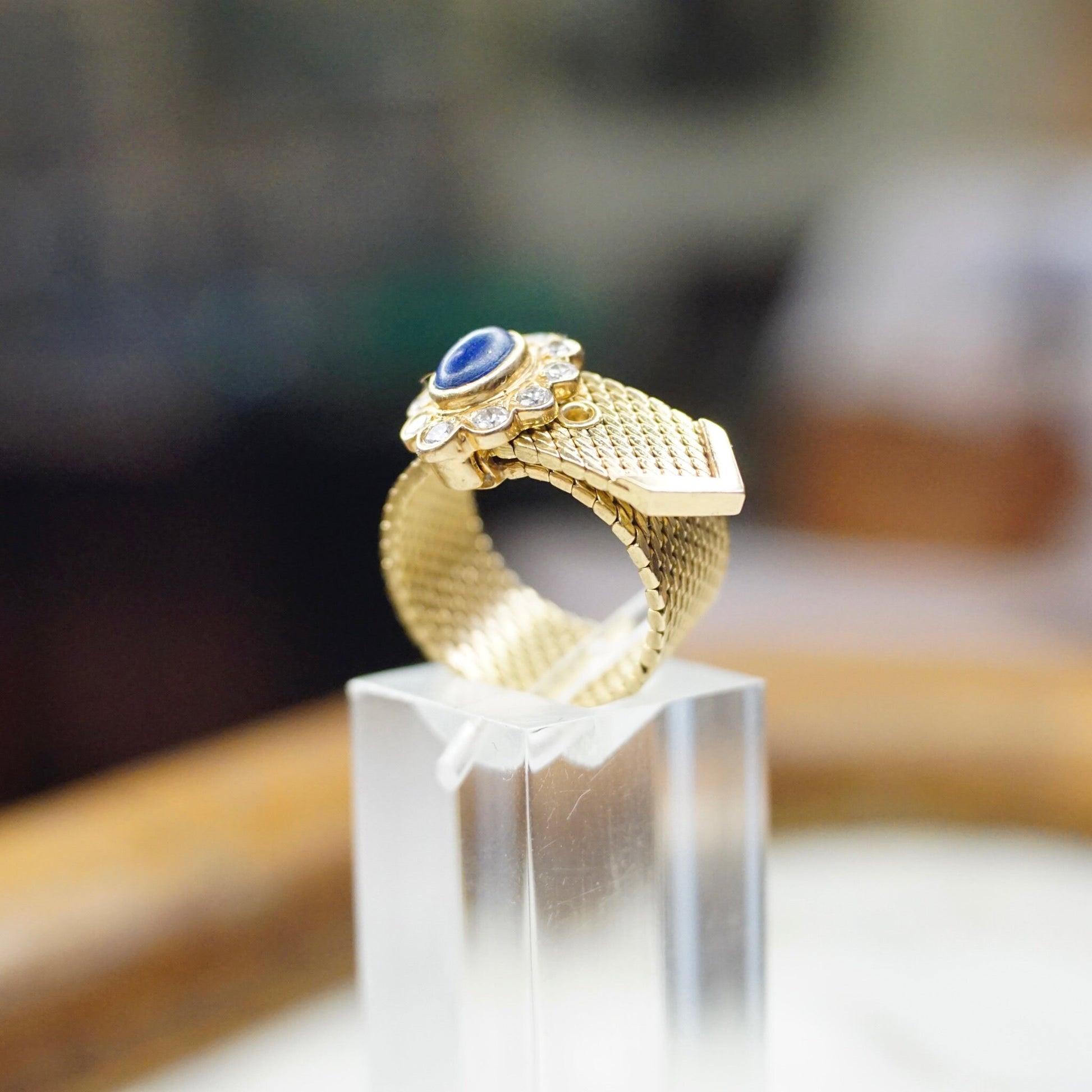 Vintage 14K yellow gold adjustable mesh belt buckle ring featuring a lapis lazuli cabochon accented by a diamond-encrusted flower, against a blurred background.