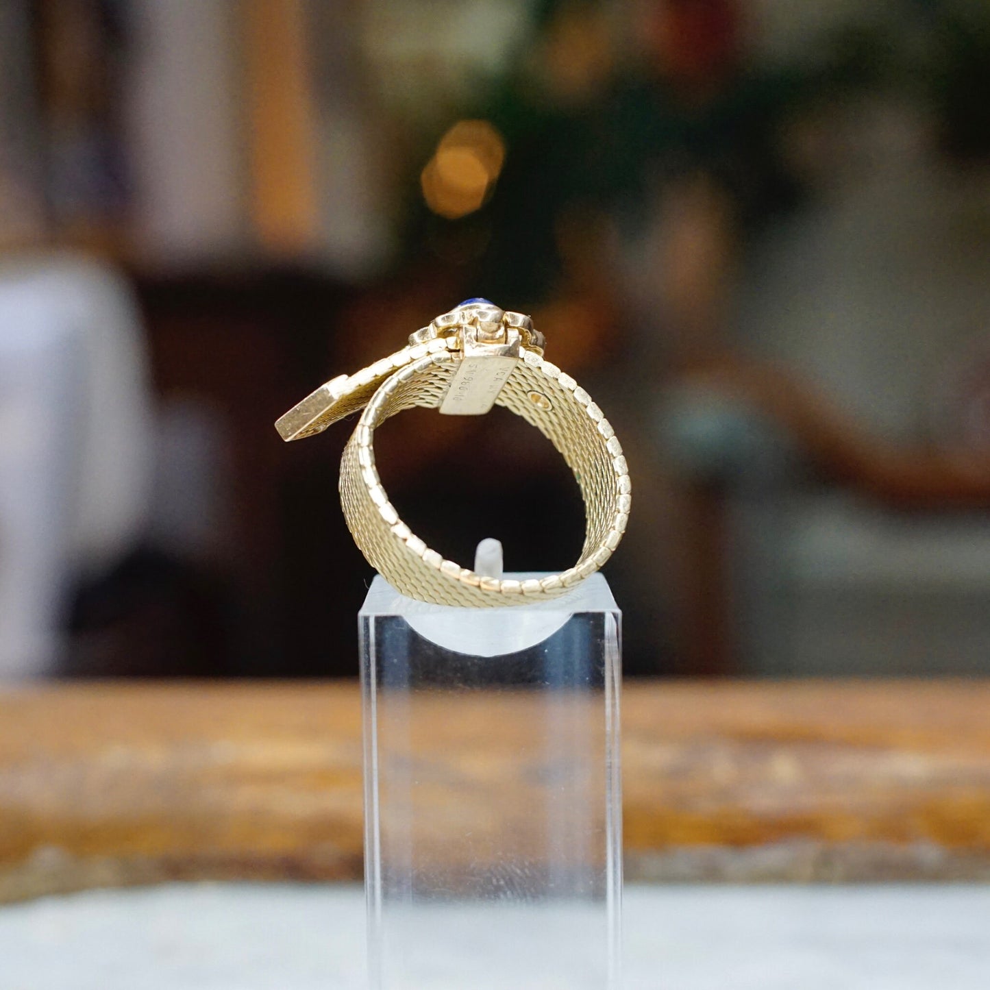 Vintage 14K gold mesh belt buckle ring with lapis lazuli and diamond flower design on clear display stand, with blurred interior background.