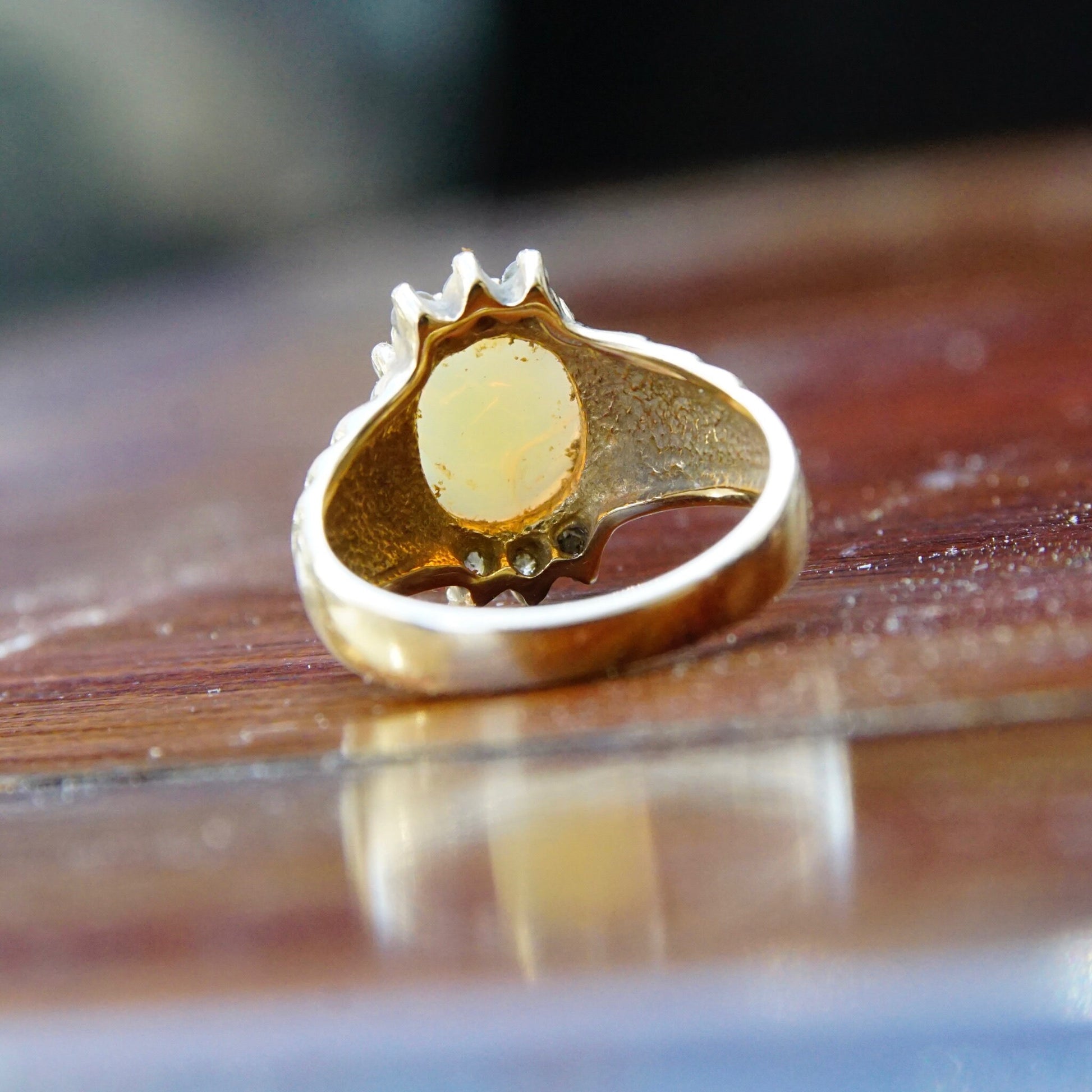 Vintage 14K yellow gold cocktail ring featuring a beautiful oval opal center stone flanked by small accent diamonds, shown on a reflective wooden surface.