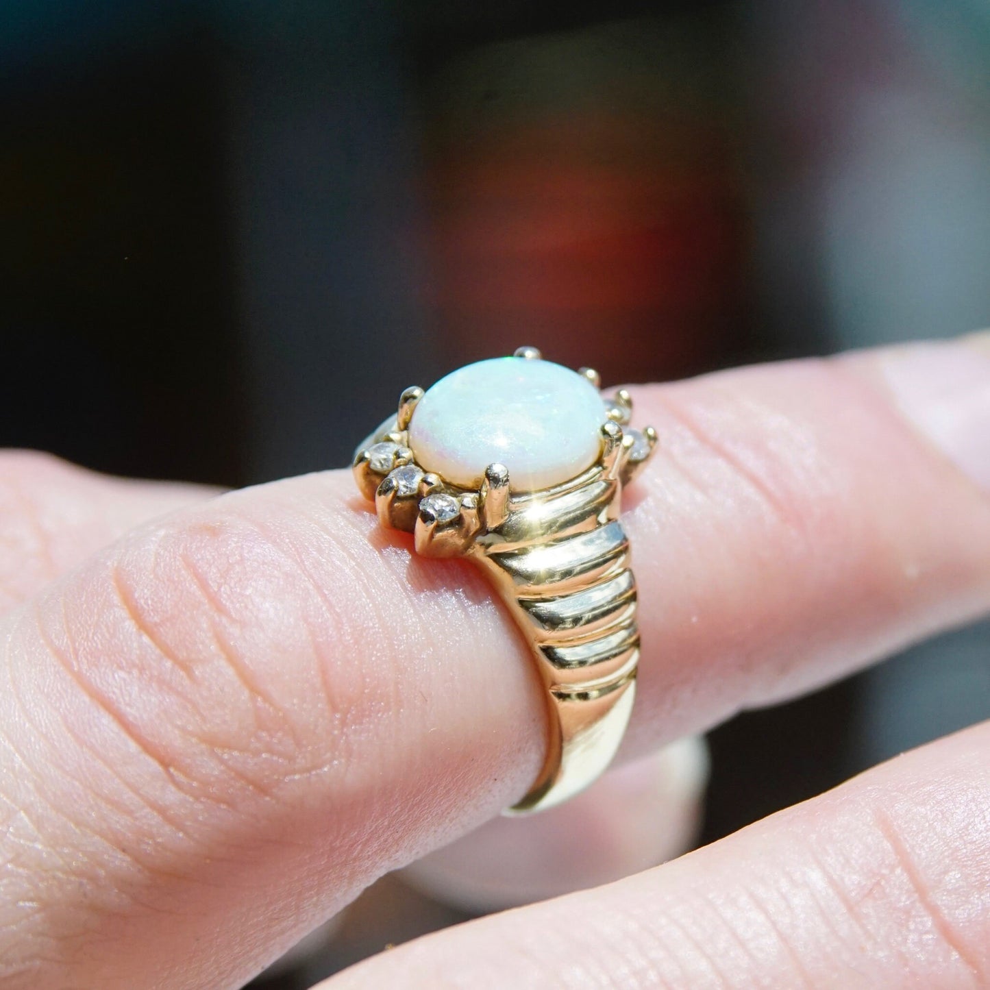 14K yellow gold vintage cocktail ring featuring a beautiful oval opal center stone accented by small diamonds, shown on a finger against a blurred background.