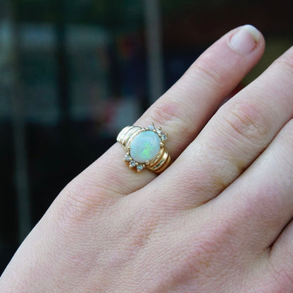 Vintage 14K gold cocktail ring featuring a beautiful center opal stone accented by tiny diamonds, shown on a woman's finger against a blurred background.