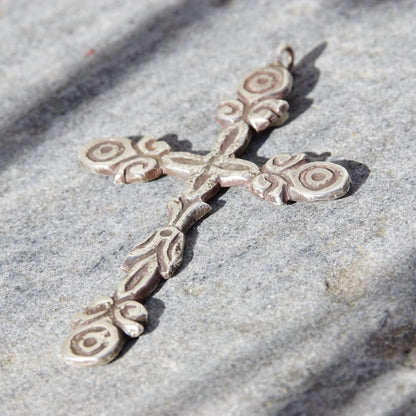 Ornate hammered sterling silver gothic cross pendant with oxidized silver design lying on gray textured fabric, creating shadows and contrast.