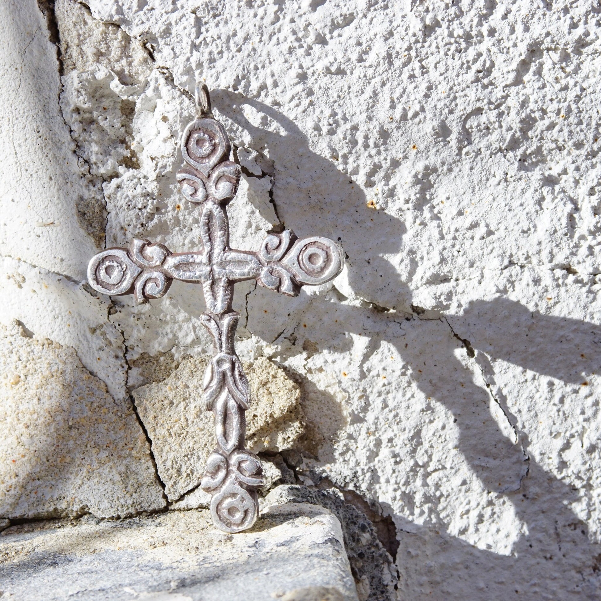 Ornate sterling silver gothic cross pendant with hammered, oxidized detailing set against a textured white stone background