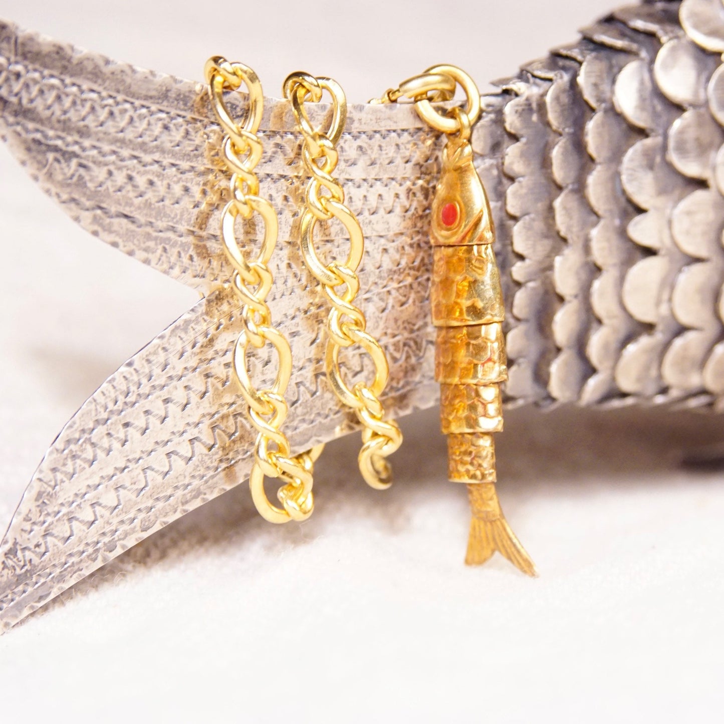 18K yellow gold articulated fish charm pendant with red cabochon eyes and movable scaled body, hanging from a silver mesh chain.