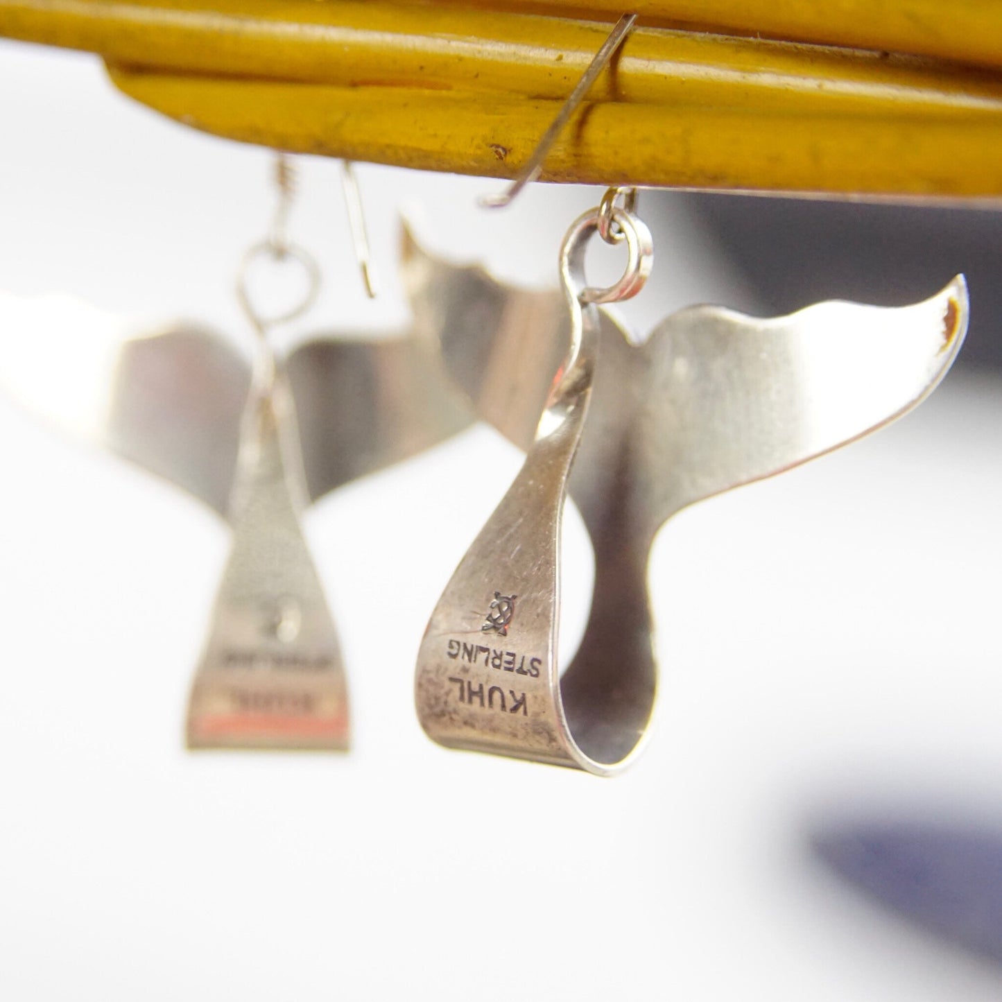 Vintage Sterling Silver Whale or Dolphin Tail Dangle Earrings hang from a yellow wooden rod, with a blurred background behind them. The minimalist silver earrings resemble the elegant curved shape of a whale or dolphin's tail on simple fish hook findings, stamped 925 to indicate their sterling silver purity.