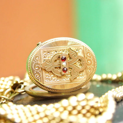 Antique gold locket necklace with intricate etched design and gemstones on a gold chain, displayed on a green background surrounded by pearls.