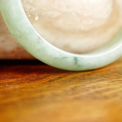 Closeup of a light green jade bangle bracelet resting on a wooden surface, showing the marbled texture and smooth, rounded shape of the solid jade bangle.