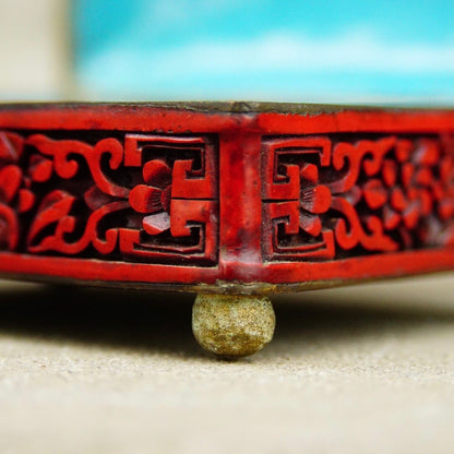 Antique Chinese carved cinnabar enamel hinge box with lid, featuring intricate figural landscape, flora and fauna motifs, seen from a low angle focusing on the carved cinnabar detail and brass ball foot, against a light colored background.