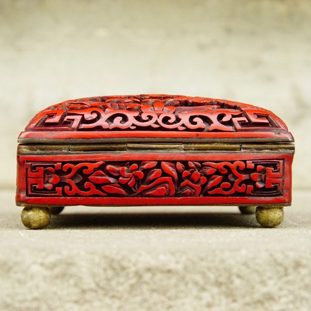Antique Chinese carved cinnabar enamel box with intricate floral and figural landscape motifs, ornate hinged lid, and brass feet, measuring 4 inches wide, 3.5 inches deep and 2 inches high.
