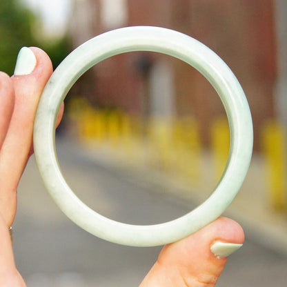 Hands holding a solid light green jade bangle bracelet outdoors, with yellow flowers blurred in the background