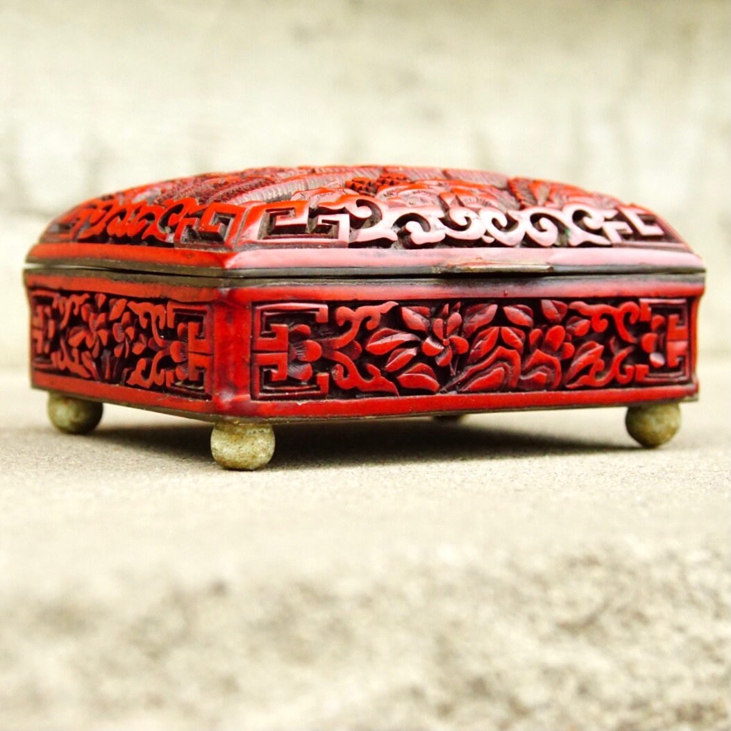 Antique Chinese carved cinnabar lacquer box with hinged lid featuring intricate figural landscape, floral and fauna motifs in red and black, measuring 4 inches wide, 3.5 inches deep and 2 inches high.