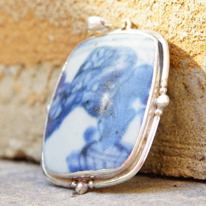 Vintage sterling silver pendant featuring inlaid blue and white ceramic pottery shard, measuring 2 3/4 inches long, set in silver frame against textured beige stone background.