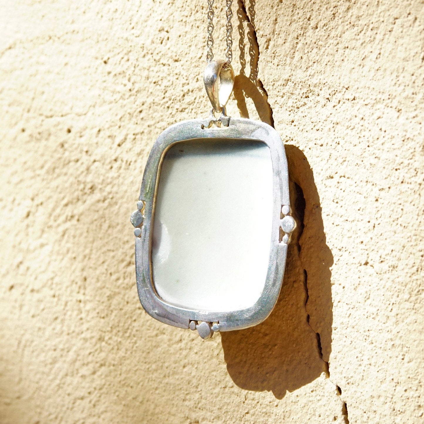 Vintage sterling silver inlaid ceramic pottery shard pendant with blue and white porcelain framed in silver, measuring 2 3/4 inches long, hanging against a textured beige wall