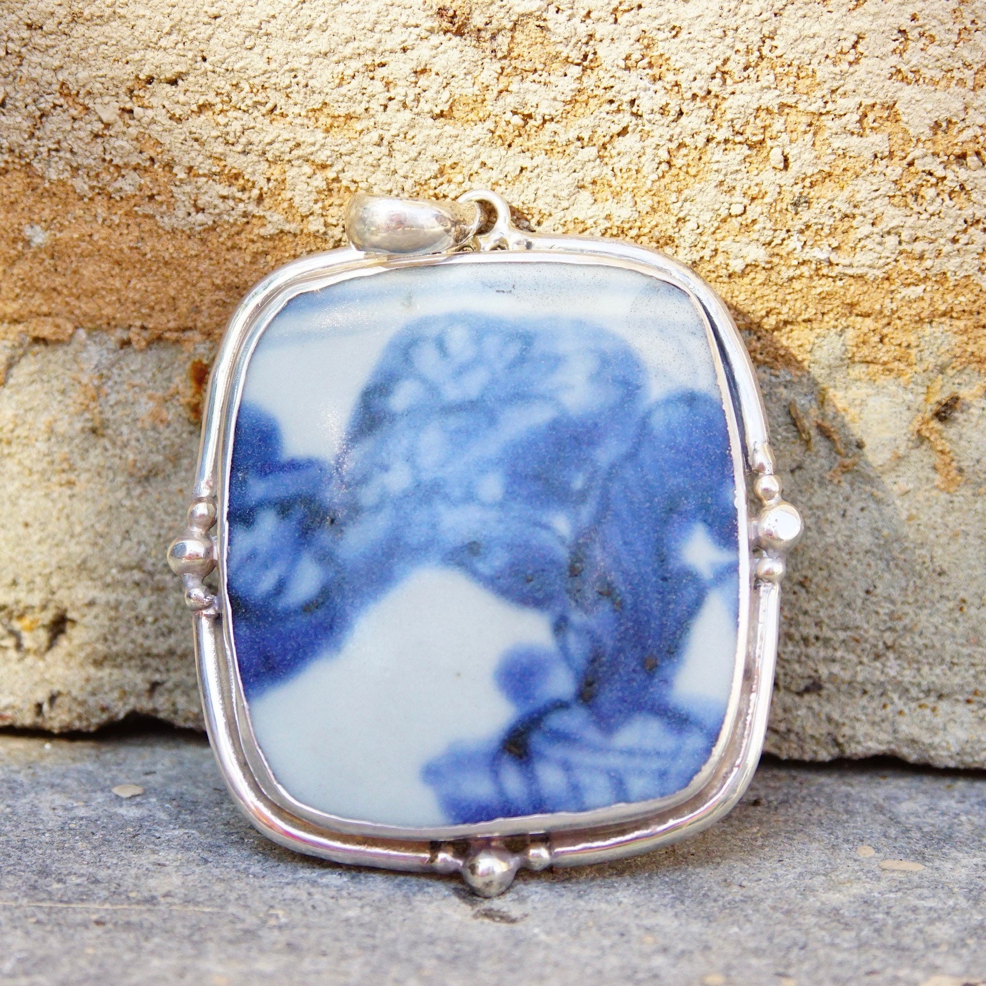 Vintage sterling silver pendant with inlaid blue and white ceramic pottery shard, measuring 2 3/4 inches long, set in a silver frame against a textured stone background.