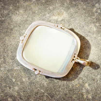 Vintage sterling silver pendant featuring an inlaid blue and white ceramic pottery shard, measuring 2 3/4 inches long, set in a silver frame against a textured stone background.