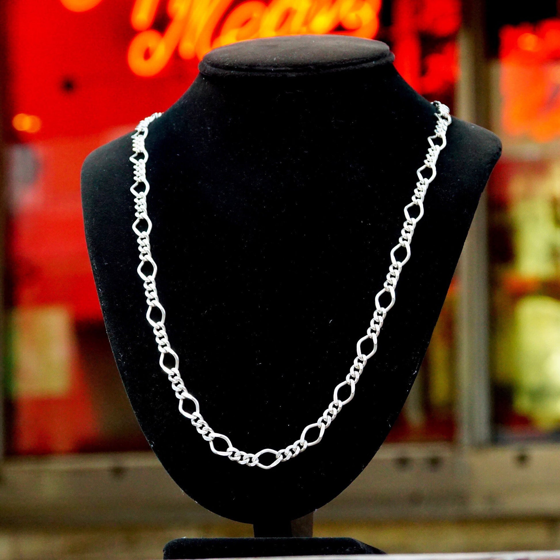 Vintage Milor 925 sterling silver figaro chain necklace with alternating 5mm curb links and 8.5mm long links on a black velvet jewelry display stand in front of a blurred red neon sign background.