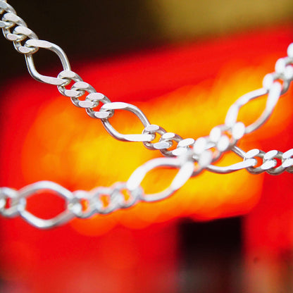 Shiny sterling silver figaro chain necklace with alternating curb and elongated oval links against blurred red and yellow background