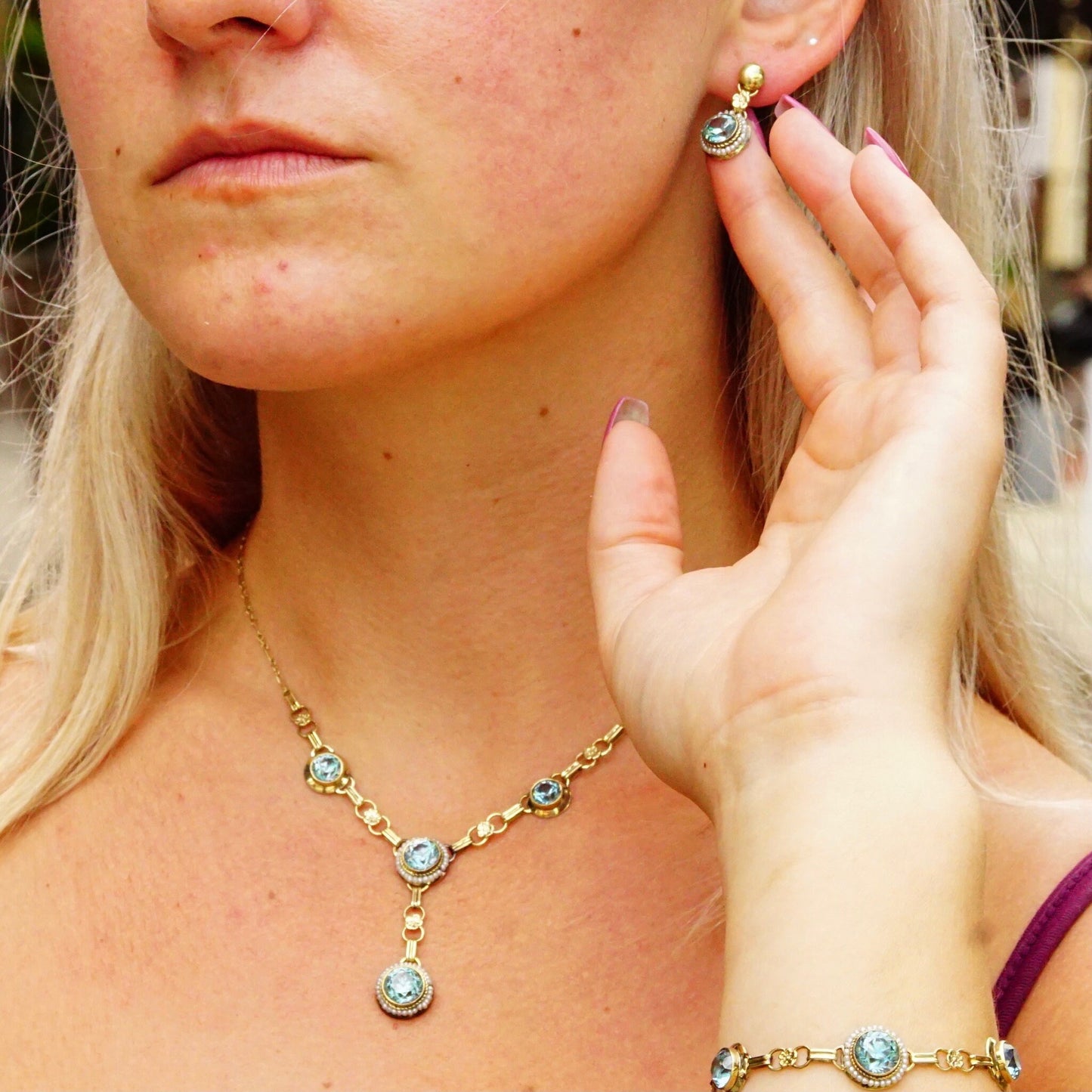 Vintage 14K gold aquamarine and seed pearl jewelry set on a young woman, including a Y-pendant necklace, bracelet, screw back earrings, and brooch with brilliant blue gemstones, shown on her neck, wrist and touching her chin.