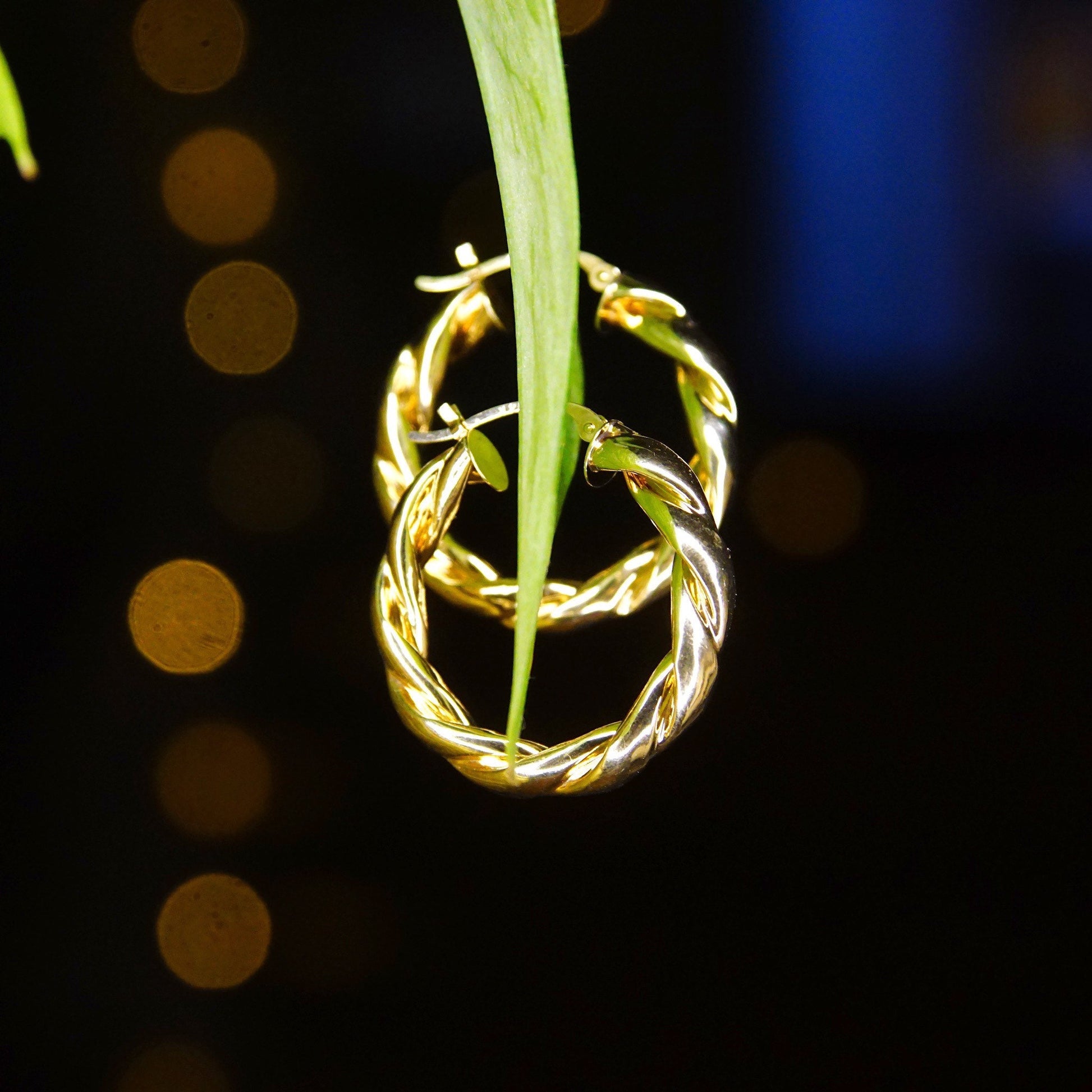 Vintage 14K yellow gold twisted hoop earrings hanging from green leaf, illuminated against blurred dark background with golden bokeh circles