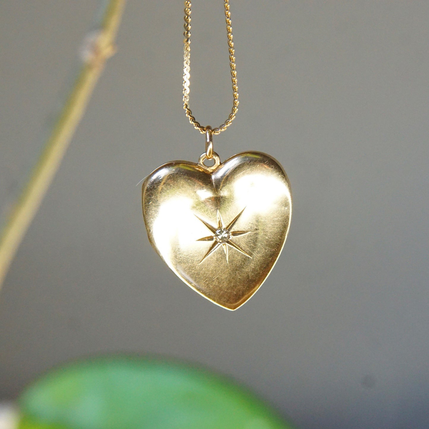 Antique 14K gold heart locket necklace with diamond starburst design, hanging from a thin gold chain against a blurred green background.