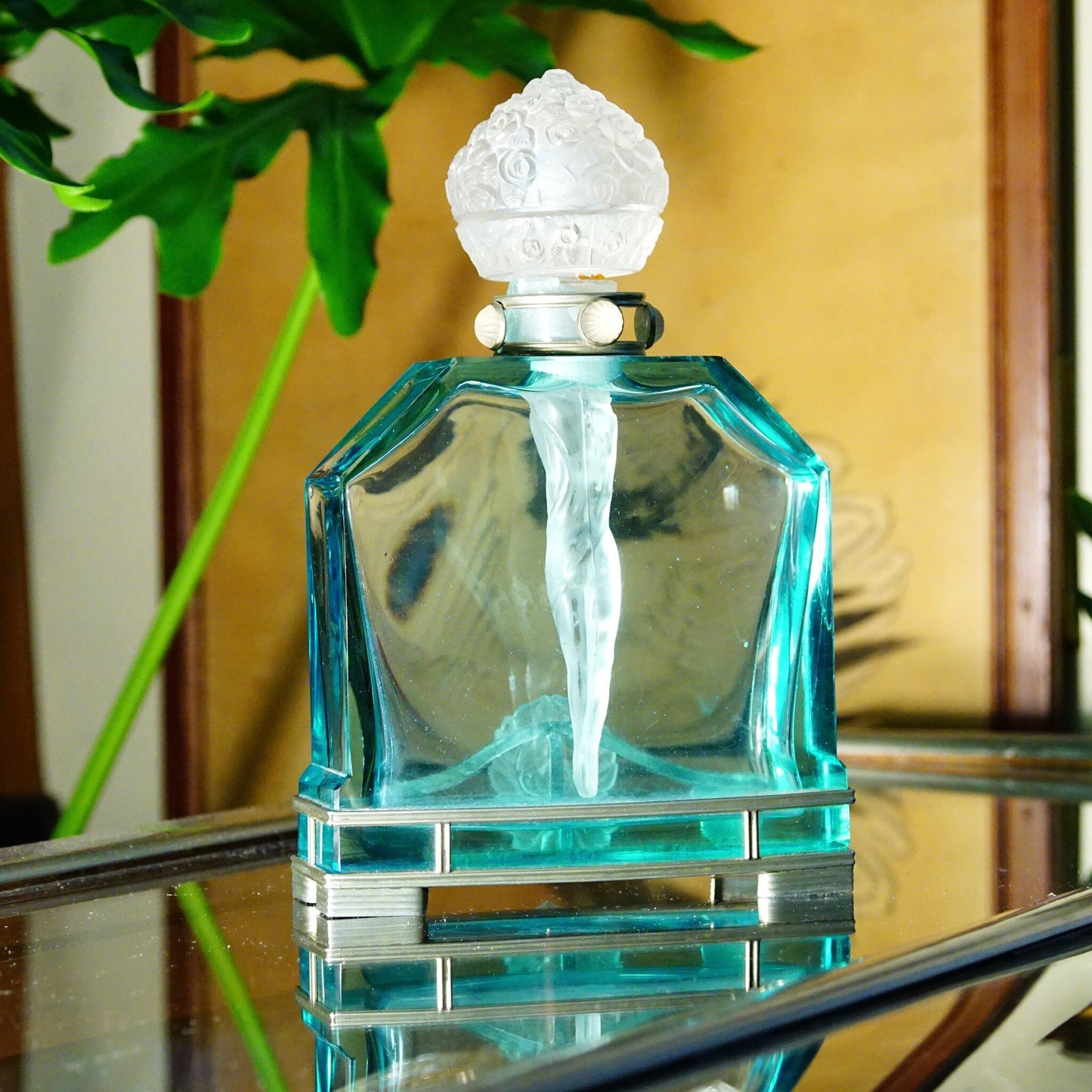 Antique 1920s Austrian blue crystal perfume bottle with frosted glass nude figure dauber and carved glass stopper on mirrored tray with plant in background.