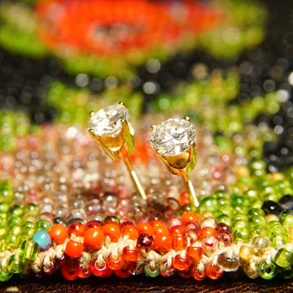 Two diamond stud earrings with sparkly round-cut diamonds set in yellow gold prongs, displayed on a colorful background of orange, red, green and black beads.