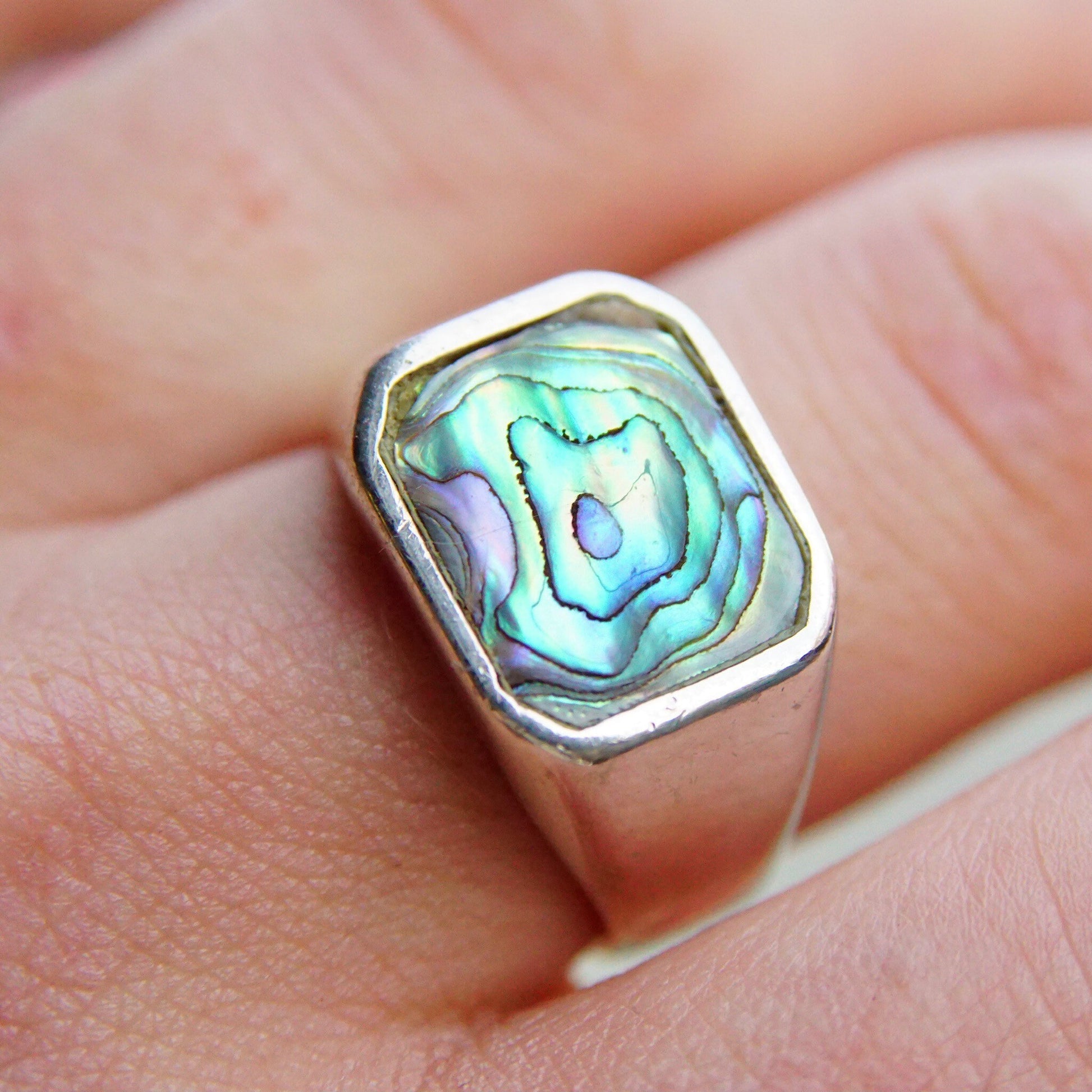 Vintage sterling silver signet ring with iridescent blue-green abalone shell inlay, depicting an abstract swirling pattern. The unisex ring is shown on a person's finger against a peach colored fabric background.