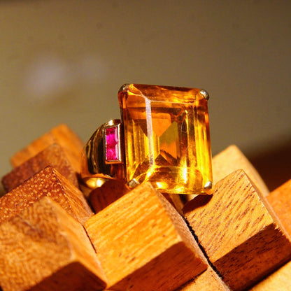 Large emerald-cut citrine gemstone cocktail ring set in 14K yellow gold with ruby accents, displayed on wooden blocks, estate jewelry.