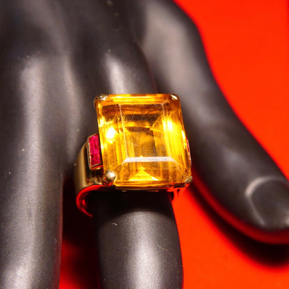 14K yellow gold cocktail ring featuring a large emerald-cut citrine gemstone accented by ruby, displayed on a mannequin hand against a bright red background.