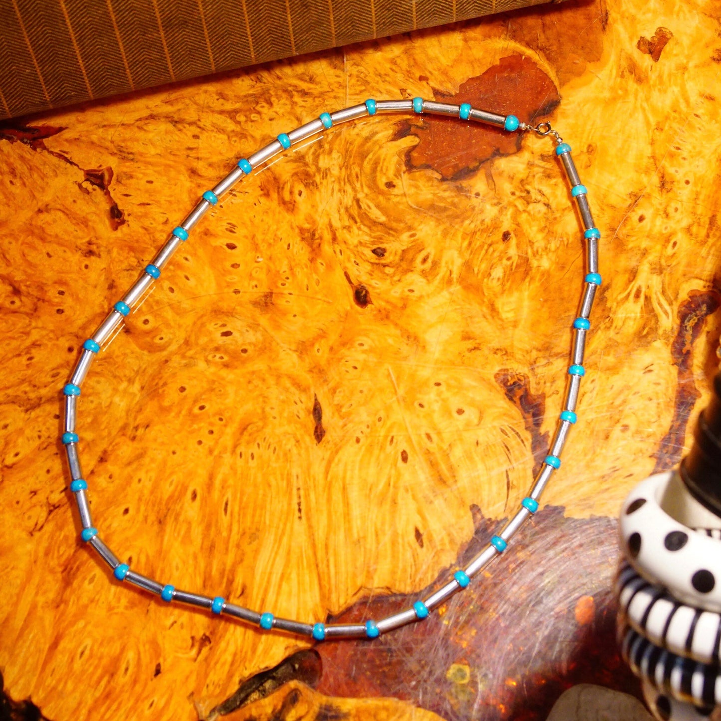 Vintage Navajo turquoise pearl bench barrel bead necklace with sterling silver tube beads and blue turquoise spacers, laying on an orange patterned textile background alongside a decorative skull sculpture.